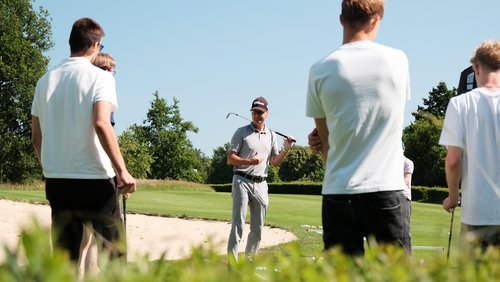 Pitchen bei Sonnenschein Ein Golftrainer steht mit einem Golfschläger vor einer Gruppe von Schülern auf einem sonnigen Golfplatz. Die Schüler hören aufmerksam zu und halten ebenfalls Schläger in der Hand. Im Hintergrund sind grüne Wiesen, Bäume und ein Sandbunker zu sehen.