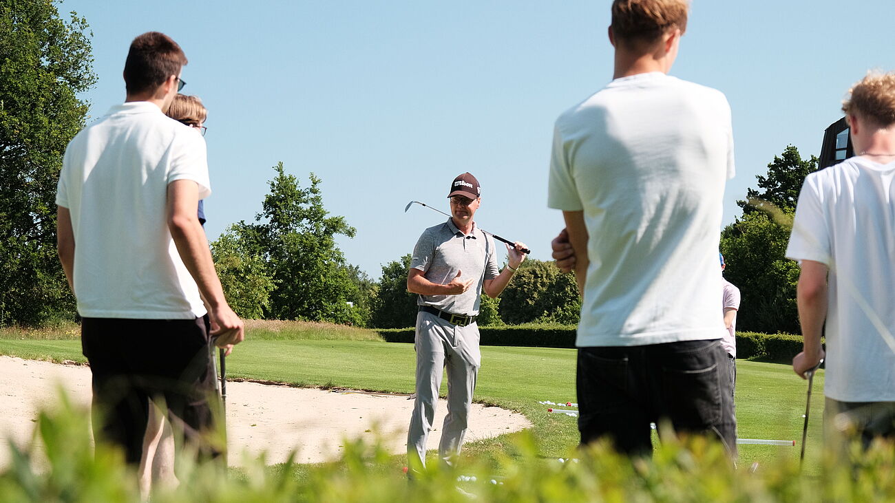 Ein Golftrainer steht mit einem Golfschläger vor einer Gruppe von Schülern auf einem sonnigen Golfplatz. Die Schüler hören aufmerksam zu und halten ebenfalls Schläger in der Hand. Im Hintergrund sind grüne Wiesen, Bäume und ein Sandbunker zu sehen.