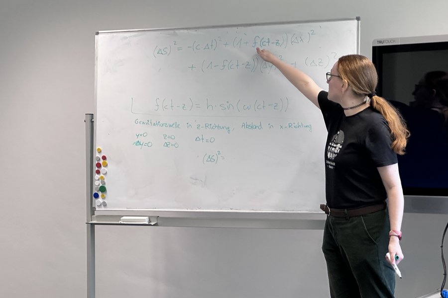 Das Bild zeigt eine Person mit langen Haaren, die seitlich zur Kamera steht und auf ein Whiteboard neben sich zeigt. Das Whiteboard ist mit wissenschaftlichen Formeln beschrieben
