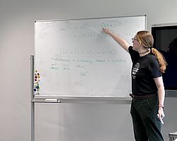 Das Bild zeigt eine Person mit langen Haaren, die seitlich zur Kamera steht und auf ein Whiteboard neben sich zeigt. Das Whiteboard ist mit wissenschaftlichen Formeln beschrieben